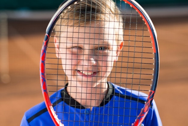 Niño cubriéndose la cara con una raqueta de tenis.