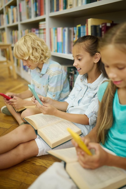 Niño y dos niñas mirando teléfonos inteligentes en la biblioteca