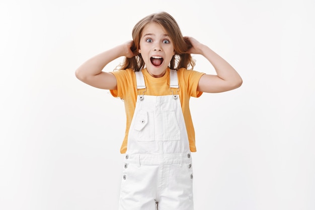 Niño emocionado feliz divirtiéndose, de pie juguetón y sorprendido, niña tocando el cabello levantando el corte de pelo en el aire, gritando divertido y alegre, expresando un estado de ánimo alegre entusiasta, pared blanca de pie