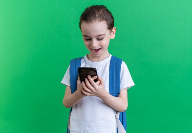 Niño emocionado con mochila sosteniendo y mirando el teléfono móvil aislado en la pared verde con espacio de copia