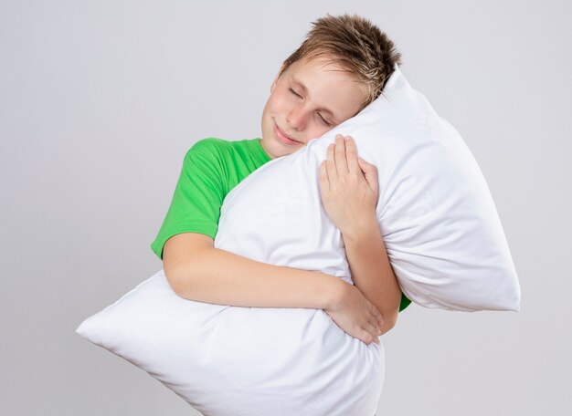 Niño enfermo en camiseta verde abrazando la almohada con los ojos cerrados sonriendo de pie sobre la pared blanca