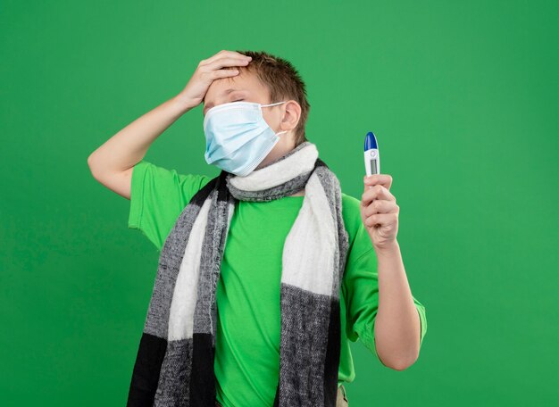 Niño enfermo en camiseta verde y bufanda cálida alrededor de su cuello con máscara protectora facial sosteniendo un triómetro que parece confundido y muy ansioso con la mano en la cabeza de pie sobre la pared verde