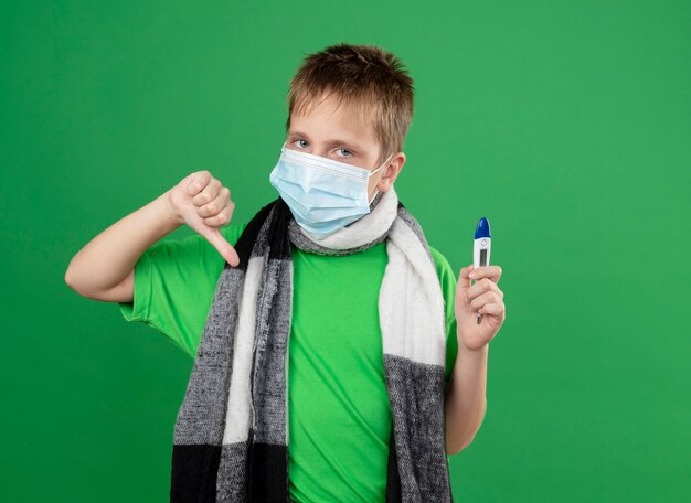 Niño enfermo en camiseta verde y bufanda cálida alrededor de su cuello con máscara protectora facial sosteniendo el triómetro sintiéndose bien mostrando los pulgares hacia abajo de pie sobre fondo verde