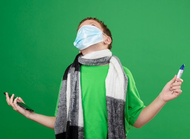 Niño enfermo en camiseta verde y bufanda cálida alrededor de su cuello con máscara protectora facial sosteniendo un triómetro y un teléfono inteligente que parece frustrado de pie sobre fondo verde