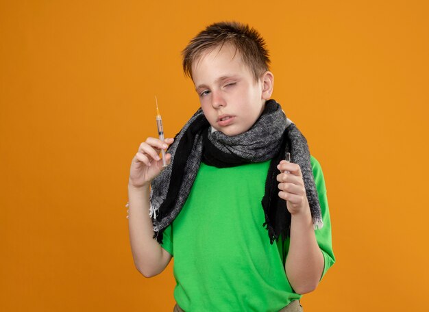 Niño enfermo en camiseta verde y bufanda cálida sensación de malestar sosteniendo ampolla y jeringa infeliz y enfermo de pie sobre la pared naranja