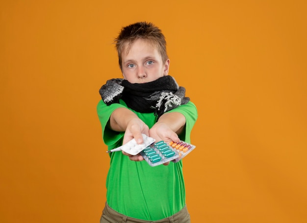 Foto gratuita niño enfermo en camiseta verde y bufanda caliente alrededor de su cuello y boca mostrando pastillas con aspecto preocupado de pie sobre la pared naranja