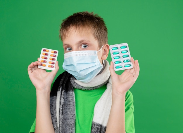 Niño enfermo en camiseta verde y bufanda caliente alrededor de su cuello con máscara protectora facial mostrando pastillas mirando a la cámara preocupado de pie sobre fondo verde