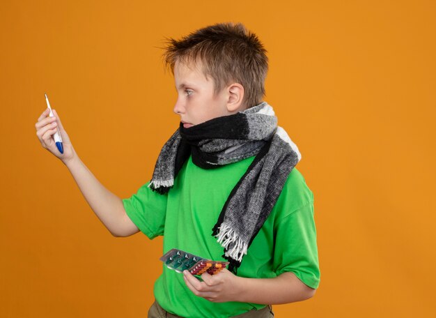 Foto gratuita niño enfermo en camiseta verde y bufanda caliente alrededor de su cuello sosteniendo píldoras mirándolos confundirse de pie sobre la pared naranja