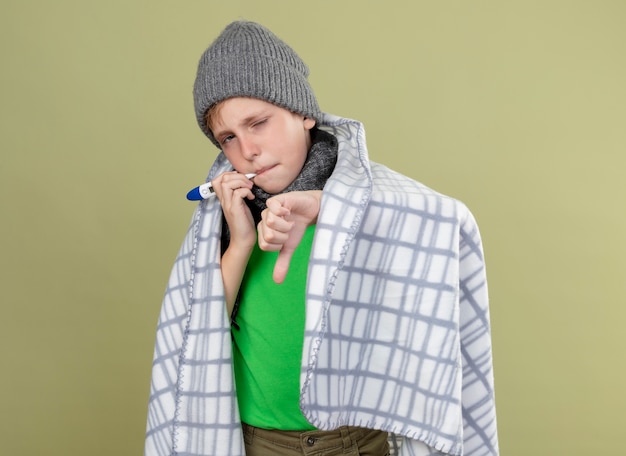 Niño enfermo con camiseta verde con bufanda caliente y sombrero envuelto en una manta poniendo termómetro en la boca midiendo la temperatura mostrando los pulgares hacia abajo de pie sobre una pared de luz