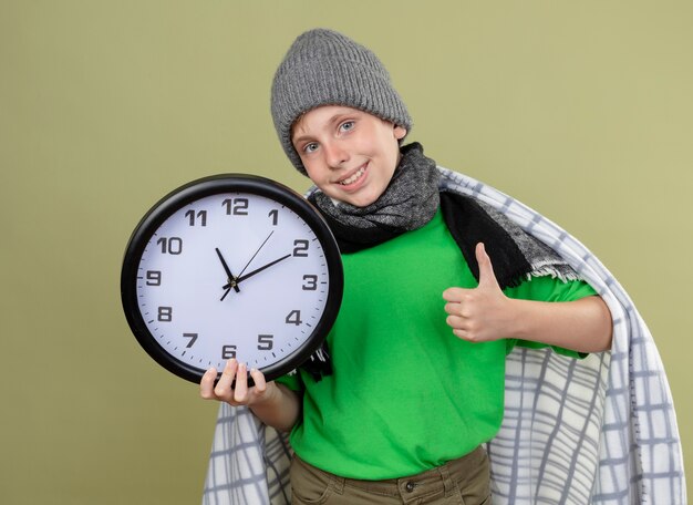 Niño enfermo con camiseta verde con bufanda caliente y sombrero envuelto en una manta con reloj de pared sonriendo mostrando los pulgares hacia arriba sintiéndose mejor parado sobre una pared de luz