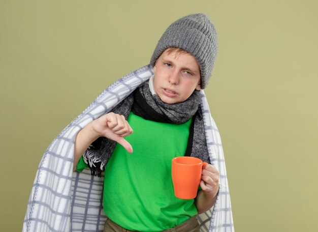 Niño enfermo con camiseta verde con bufanda caliente y sombrero envuelto en una manta sosteniendo una taza de té caliente mostrando los pulgares hacia abajo, infeliz y enfermo de pie sobre una pared de luz