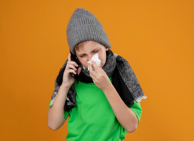 Foto gratuita niño enfermo con camiseta verde con bufanda caliente y sombrero sintiéndose mal hablando por teléfono móvil secándose la nariz con una servilleta de papel de pie sobre una pared naranja