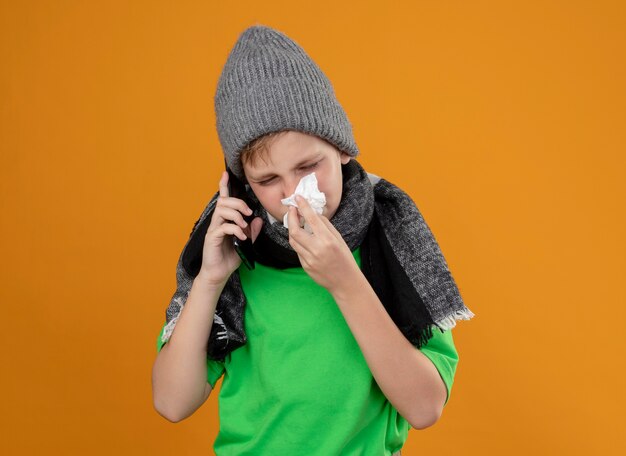 Niño enfermo con camiseta verde con bufanda caliente y sombrero sintiéndose mal hablando por teléfono móvil secándose la nariz con una servilleta de papel de pie sobre una pared naranja