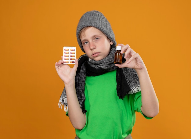 Foto gratuita niño enfermo con camiseta verde con bufanda caliente y sombrero sintiéndose mal mostrando frasco de medicina y pastillas infelices y enfermas de pie sobre la pared naranja