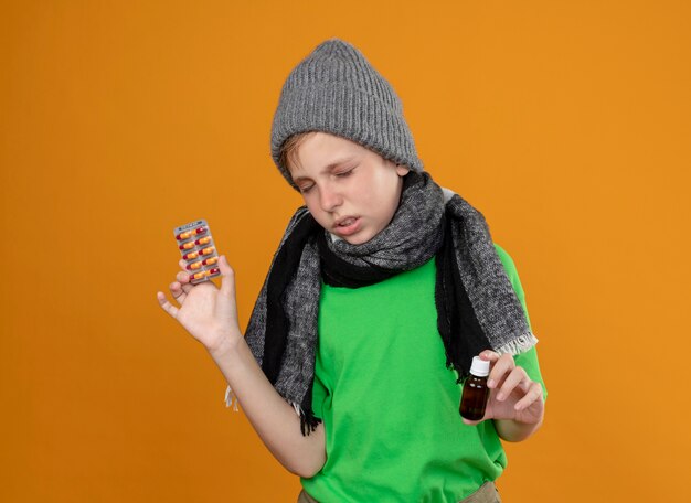 Niño enfermo con camiseta verde con bufanda caliente y sombrero sintiéndose mal sosteniendo un frasco de medicina y pastillas de pie enfermo e infeliz sobre la pared naranja