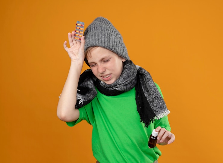 Foto gratuito niño enfermo con camiseta verde con bufanda caliente y sombrero sintiéndose mal sosteniendo un frasco de medicina y pastillas que sufren de dolor de cabeza enfermo e infeliz de pie sobre una pared naranja