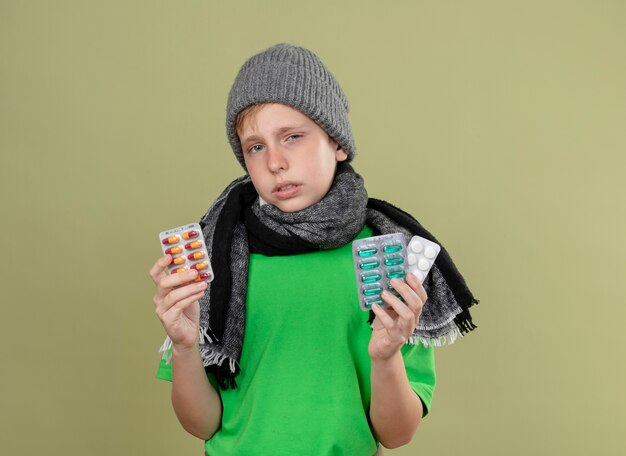 Niño enfermo con camiseta verde con bufanda caliente y sombrero sintiéndose mal sosteniendo pastillas infeliz y enfermo de pie sobre la pared de luz