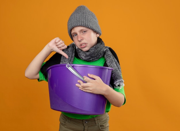 Niño enfermo con camiseta verde con bufanda caliente y sombrero sosteniendo basura sintiéndose con náuseas mostrando los pulgares hacia abajo infeliz y enfermo de pie sobre la pared naranja