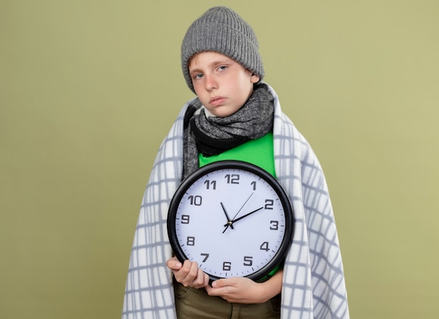 Foto gratuita niño enfermo con camiseta verde con bufanda y sombrero envuelto en una manta con reloj de pared sintiéndose mal, enfermo e infeliz de pie sobre una pared de luz