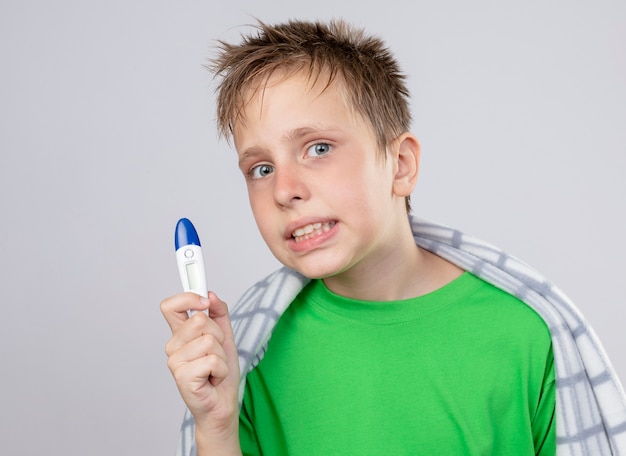 Niño enfermo en camiseta verde envuelto en una manta malestar mostrando termómetro mirando confundido parado sobre pared blanca