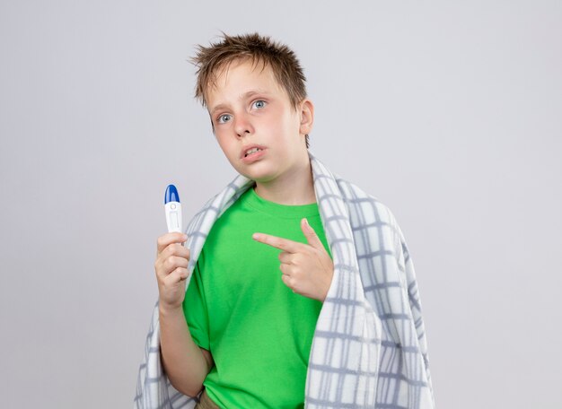 Niño enfermo en camiseta verde envuelto en una manta malestar sosteniendo el termómetro apuntando con el dedo índice a él de pie sobre la pared blanca