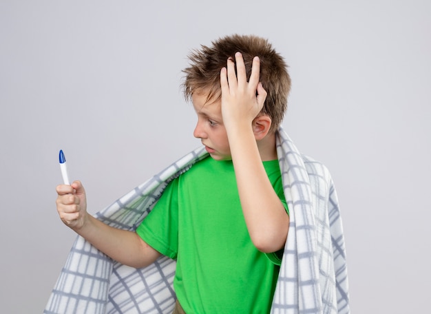 Foto gratuita niño enfermo en camiseta verde envuelto en una manta sintiéndose mal sosteniendo el termómetro mirándolo estar preocupado de pie sobre la pared blanca y confundido