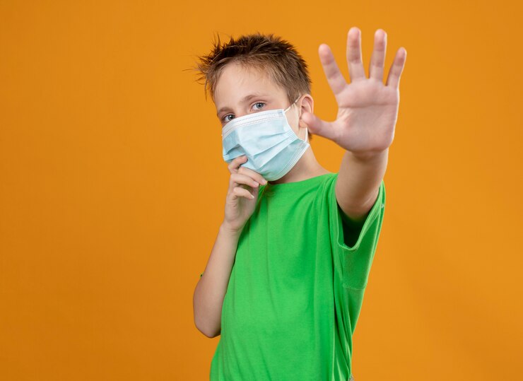 Foto gratuito niño enfermo en camiseta verde con máscara protectora facial haciendo gesto de parada con la mano de pie sobre la pared naranja