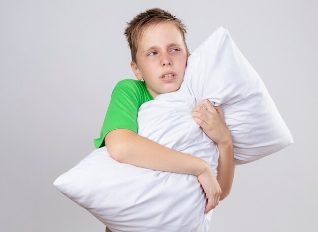 Niño enfermo en camiseta verde que se siente mal abrazando la almohada mirando a un lado infeliz y enfermo de pie sobre la pared blanca