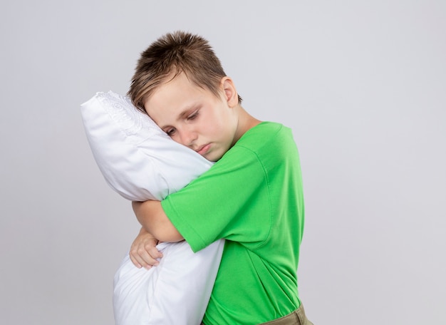 Niño enfermo en camiseta verde que se siente mal abrazando la almohada con los ojos cerrados de pie sobre la pared blanca