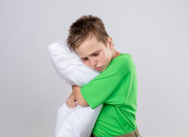 Niño enfermo en camiseta verde que se siente mal abrazando la almohada con los ojos cerrados de pie sobre la pared blanca