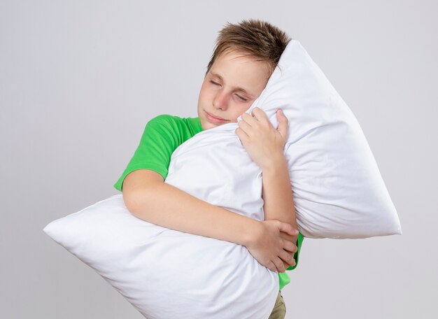 Niño enfermo en camiseta verde que se siente mal abrazando la almohada con los ojos cerrados de pie sobre la pared blanca