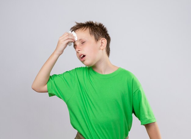Niño enfermo en camiseta verde que se siente mal limpiando su rorehead con una servilleta de papel de pie sobre una pared blanca