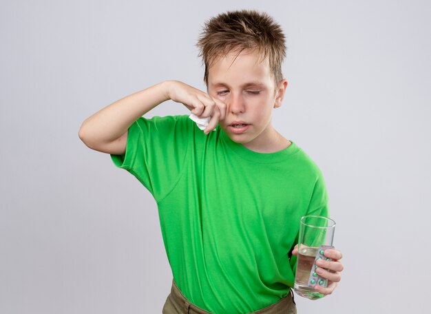 Niño enfermo en camiseta verde que se siente mal sosteniendo un vaso de agua y pastillas limpiando la nariz con una servilleta de papel que sufre de frío parado sobre una pared blanca