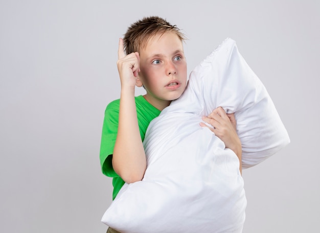 Niño enfermo en camiseta verde se siente mal abrazando la almohada mirando a un lado preocupado de pie sobre la pared blanca