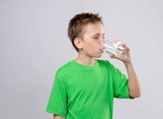 Niño enfermo en camiseta verde sintiendo agua potable indeseable de pie sobre la pared blanca