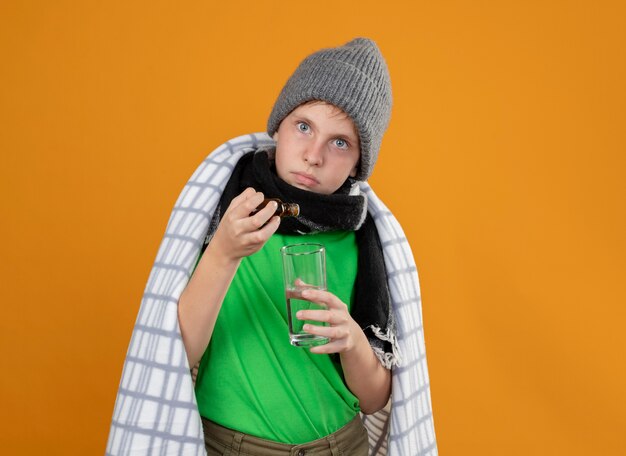 Niño enfermo con gorro y bufanda envuelto en una manta goteando gotas de la botella de medicina en un vaso de pie sobre una pared naranja