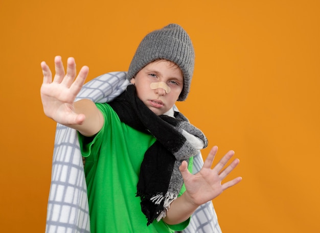 Foto gratuita niño enfermo con gorro y bufanda envuelto en una manta con parche en la nariz, infeliz y enfermo, haciendo gestos de defensa con las manos sobre la pared naranja