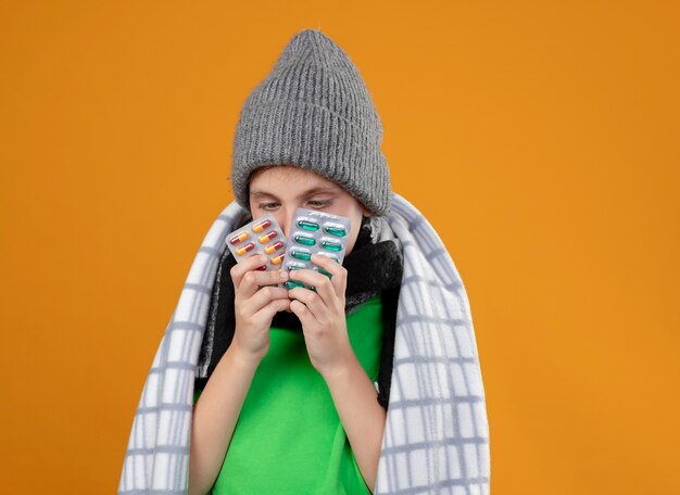 Niño enfermo con gorro y bufanda envuelto en una manta que sufre de fiebre sosteniendo pastillas sintiéndose mal parado sobre la pared naranja