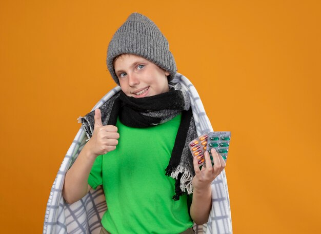 Niño enfermo con gorro y bufanda envueltos en mantas mostrando pastillas sintiéndose mejor luciendo sonriendo mostrando los pulgares para arriba de pie sobre la pared naranja