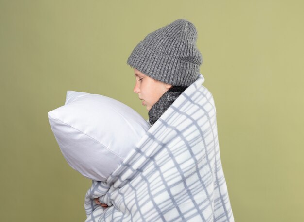 Niño enfermo con gorro envuelto en una manta sosteniendo la almohada sintiéndose mal de lado sobre la pared de luz