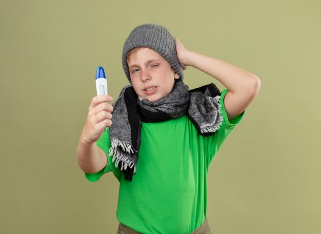 Niño enfermo vistiendo camiseta verde con bufanda caliente y sombrero sintiéndose mal mostrando termómetro confundido y disgustado de pie sobre la pared de luz