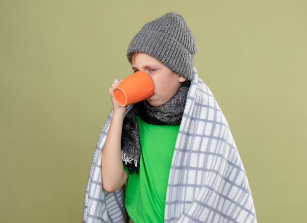 Niño enfermo vistiendo camiseta verde con bufanda y gorro envuelto en una manta bebiendo té caliente que sufre de frío de pie sobre una pared ligera