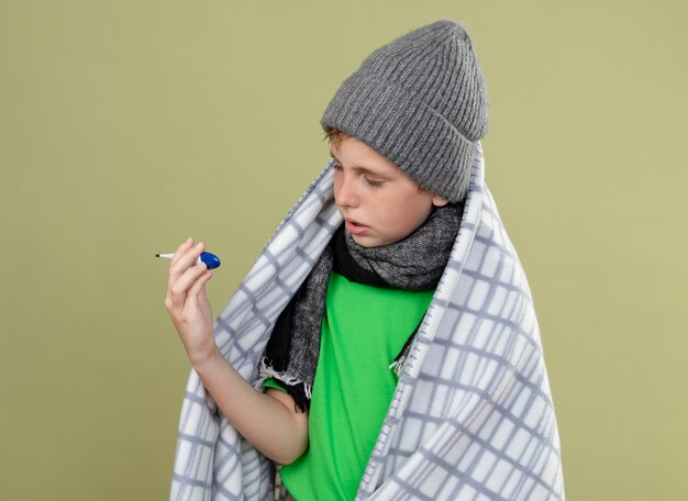 Niño enfermo vistiendo camiseta verde en cálida bufanda y sombrero envuelto en una manta sosteniendo el termómetro mirándolo preocupado de pie sobre la pared de luz