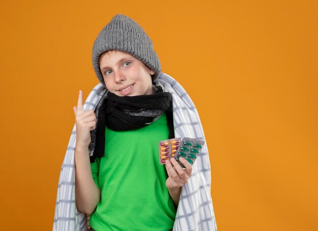 Niño enfermo vistiendo gorro y bufanda envueltos en mantas mostrando pastillas sintiéndose mejor luciendo sonriendo mostrando el dedo índice parado sobre la pared naranja