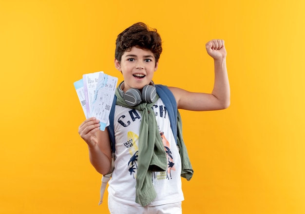 Niño de escuela alegre con bolsa trasera y auriculares sosteniendo boletos y mostrando gesto sí aislado sobre fondo amarillo