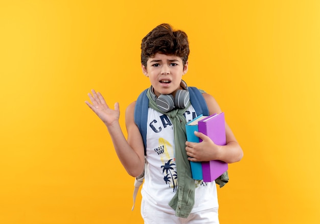 Niño de escuela confundido con mochila y auriculares sosteniendo libros y puntos con la mano al lado aislado sobre fondo amarillo con espacio de copia