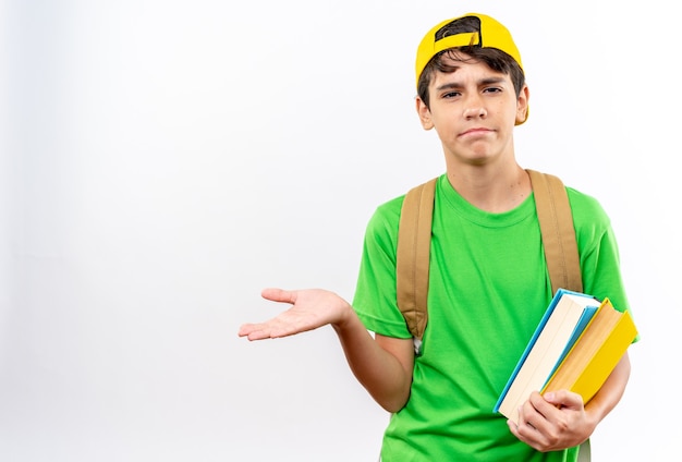 Niño de escuela joven confundido con mochila con gorra sosteniendo libros extendiendo la mano