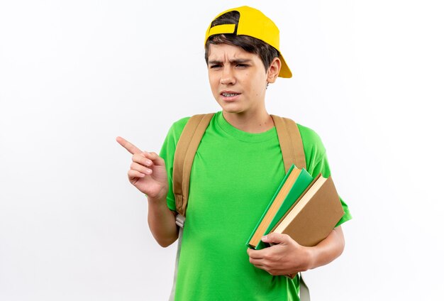 Niño de escuela joven disgustado con mochila con gorra sosteniendo libros puntos al lado