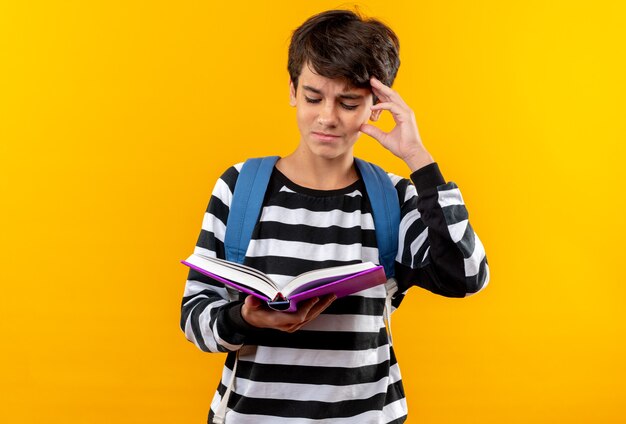 Niño de escuela joven disgustado con mochila sosteniendo y leyendo el libro poniendo la mano en la cabeza