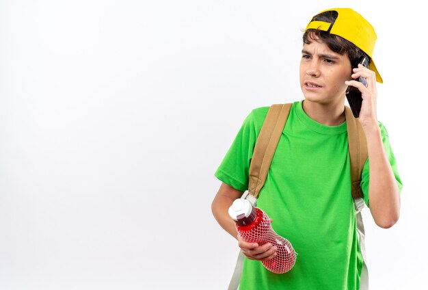 Niño de escuela joven de lado mirando disgustado con mochila con tapa sosteniendo una botella de agua habla por teléfono